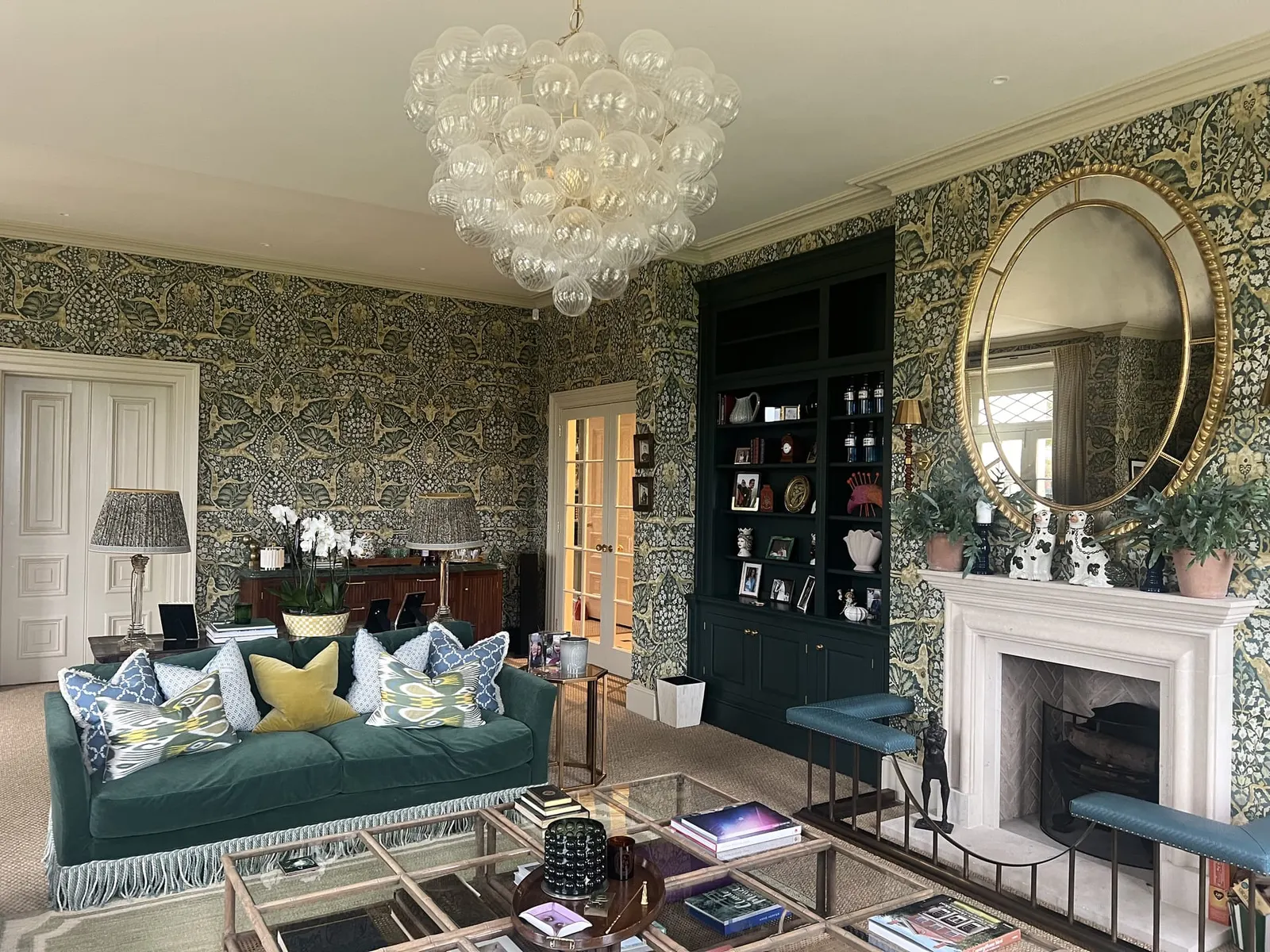 A drawing room papered in a green foliage print, with a teal velvet sofa, marble chimneypiece and an oval gilt mirror.