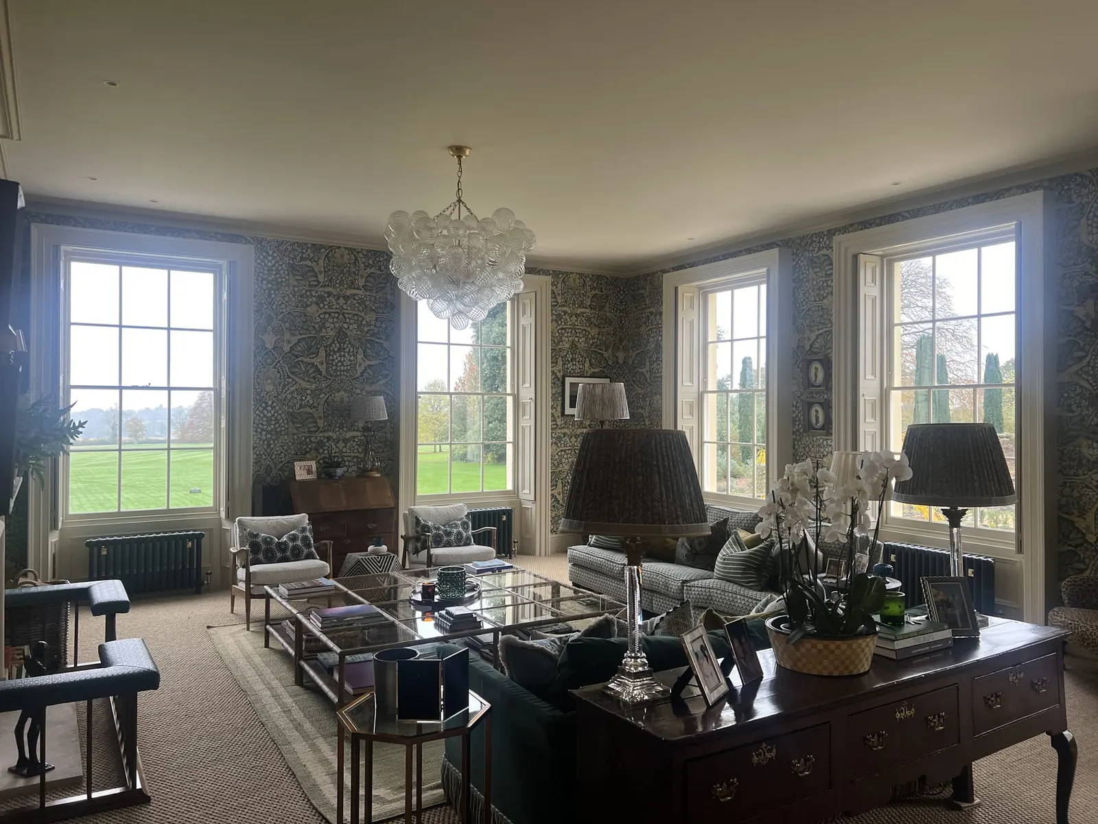A sitting room with three windows onto parkland, dark patterned walls and a glass chandelier.