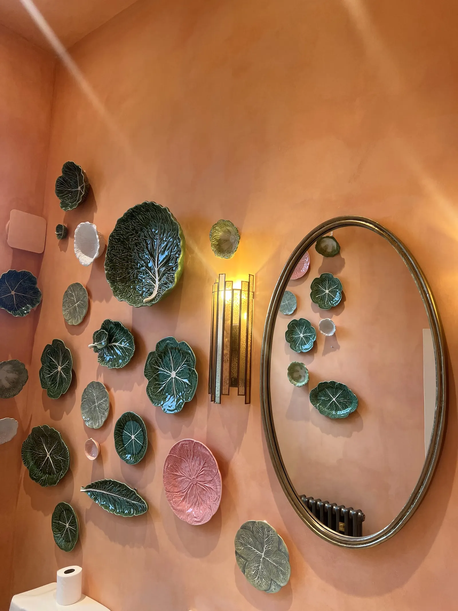 A coral-painted wall hung with green cabbage-leaf plates around an oval mirror.