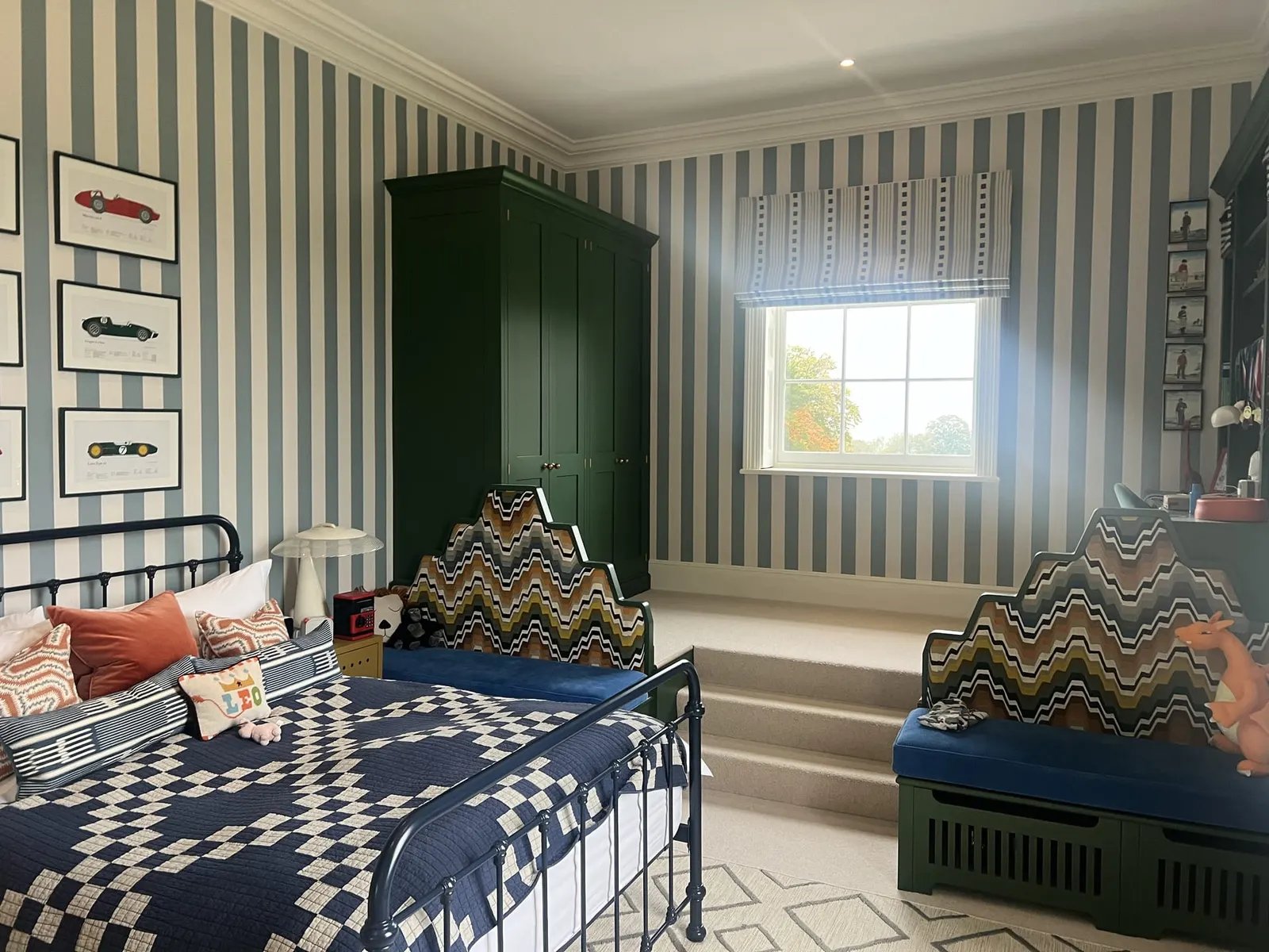 A striped bedroom with a dark green wardrobe, iron bed and a cushioned window seat.