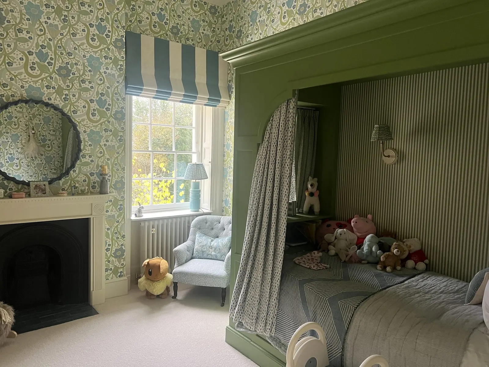 A child’s room with a green painted alcove bed under a striped canopy, beside a fireplace.