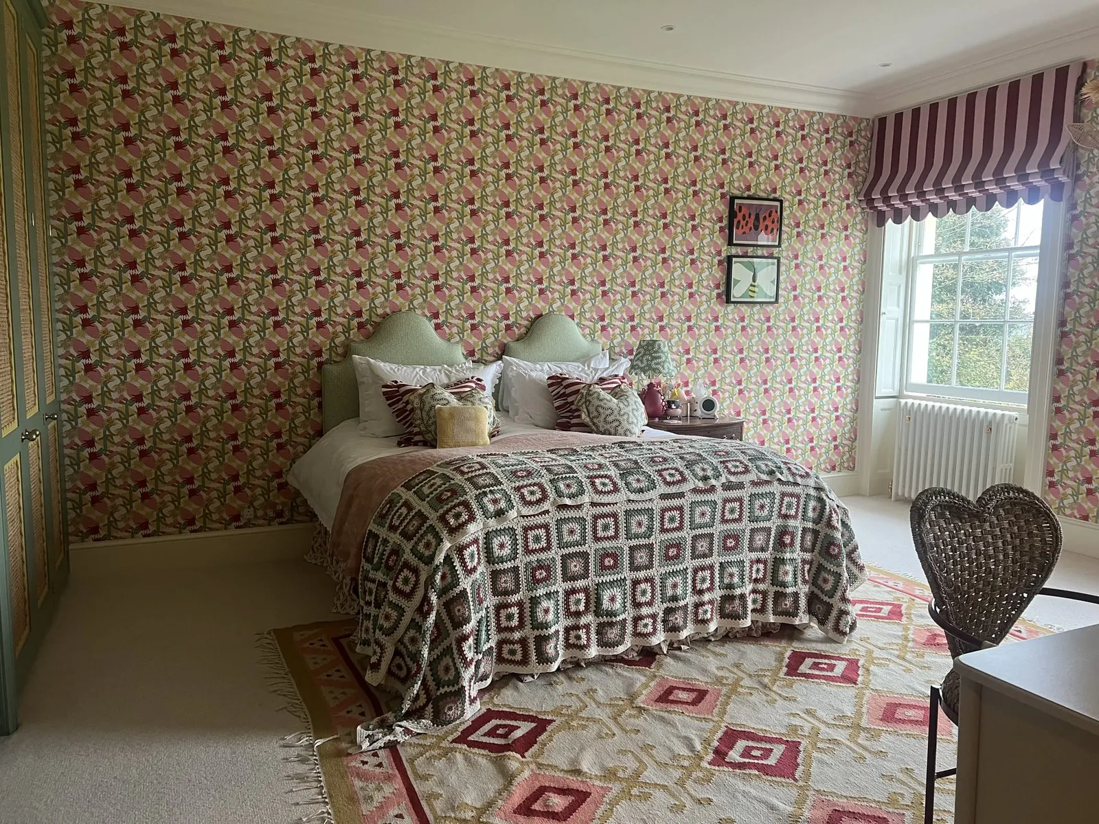 A bedroom papered in a pink floral print, with a striped blind and a patterned bedcover.