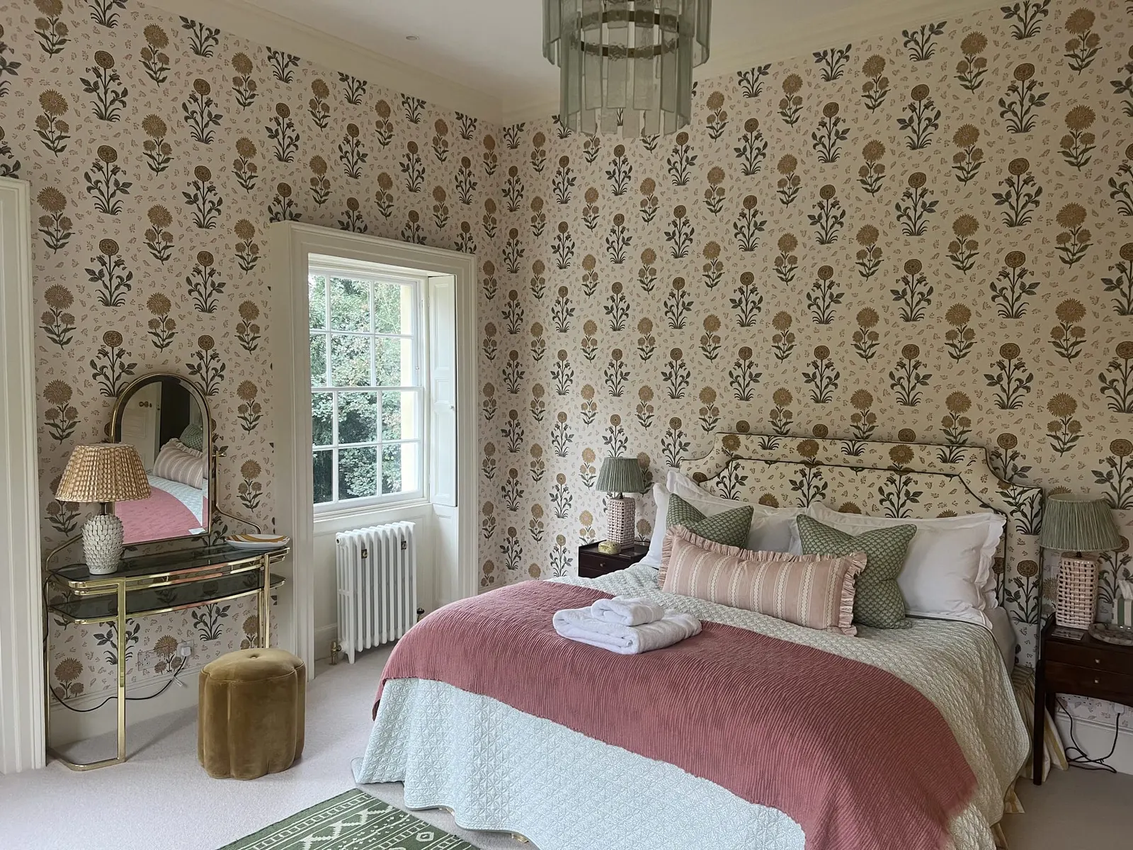 A bedroom in a block-print wallpaper, with a dressing table and a pink quilted bed.