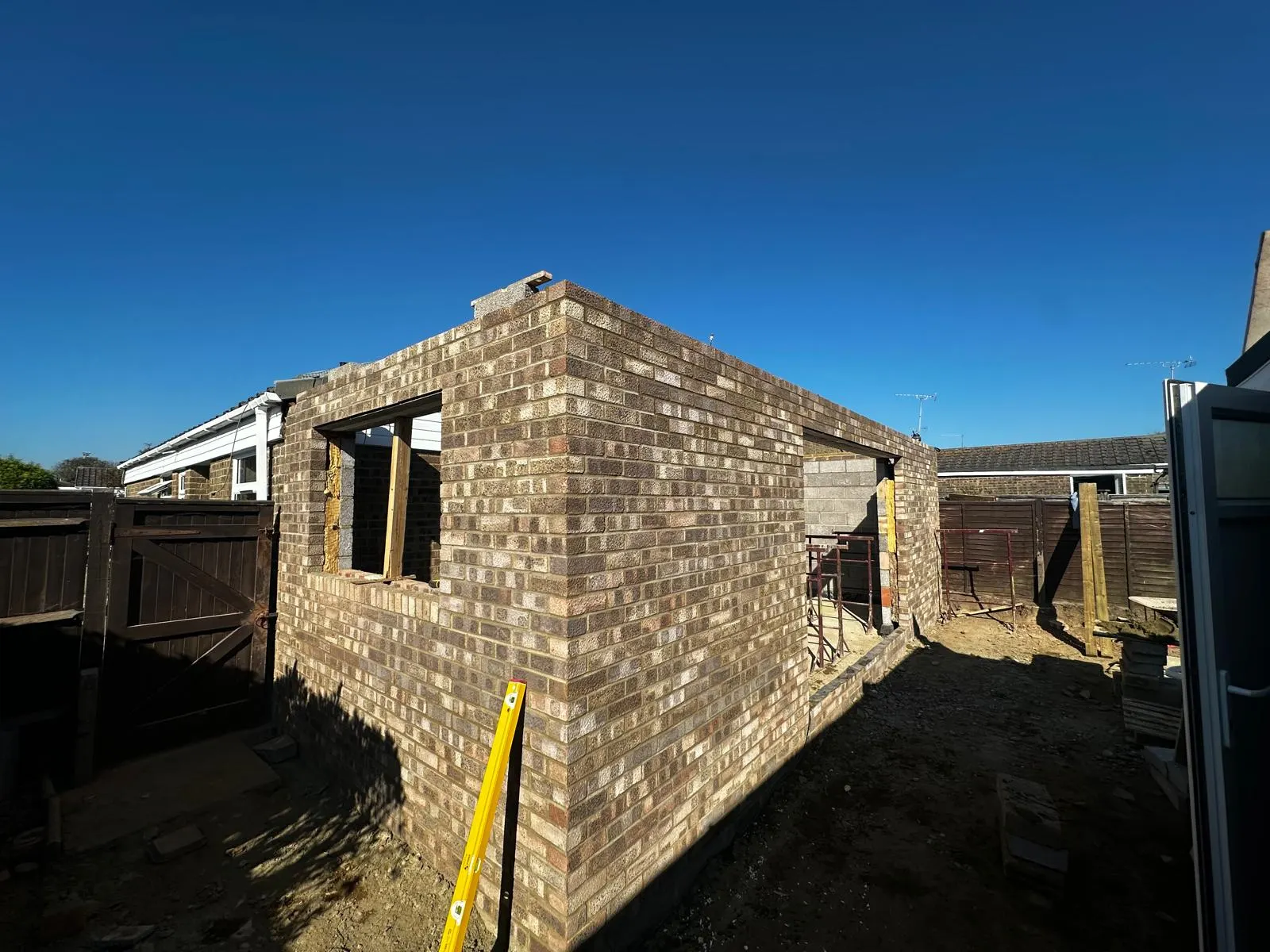 A garden room shell in buff multi brick, its openings formed, under a clear sky.