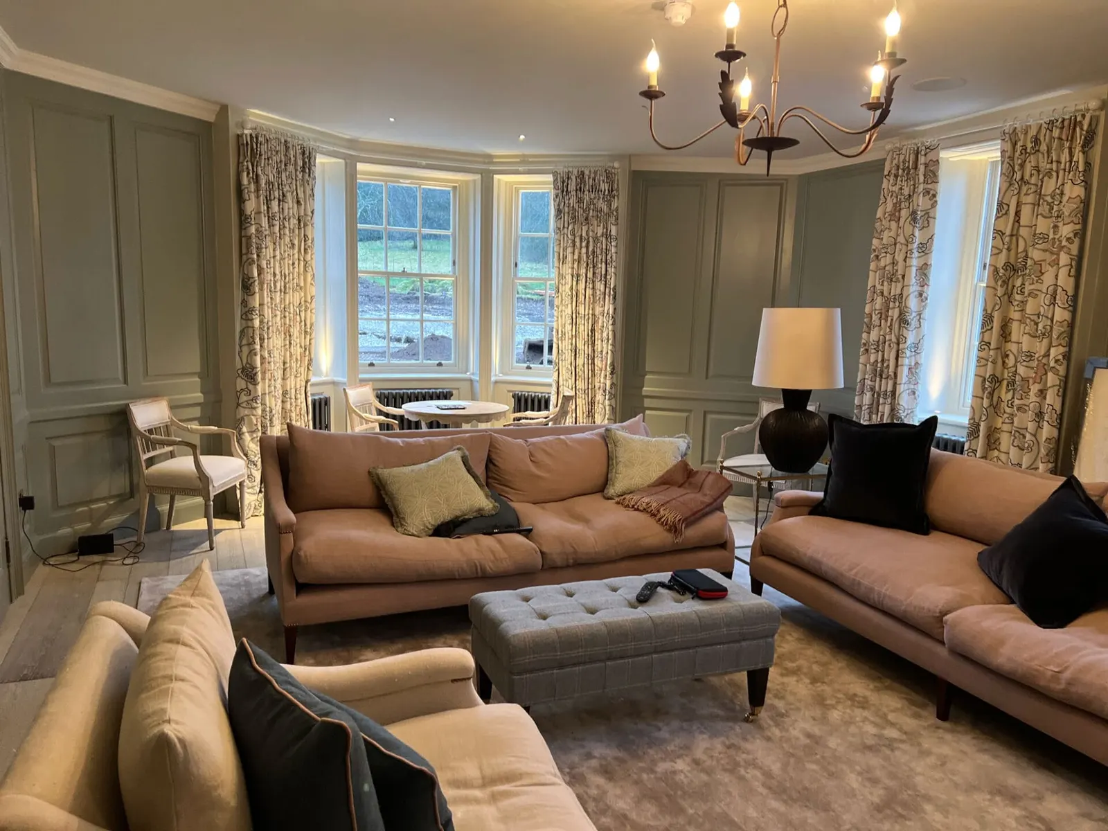 A bay-windowed sitting room with tan upholstery, a chandelier and long curtains.