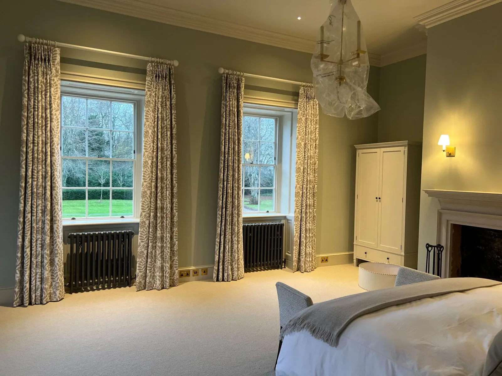 A bedroom with two curtained windows either side of the bed and a wall light above.