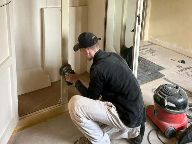 An apprentice kneeling to sand a door frame, dust extraction running beside him.